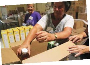 Harry Chapin Food Bank volunteers Sharon Wilmoth (L, in purple) and Lynn Madore (R, in light gray) pack boxes of food for seniors in need.
